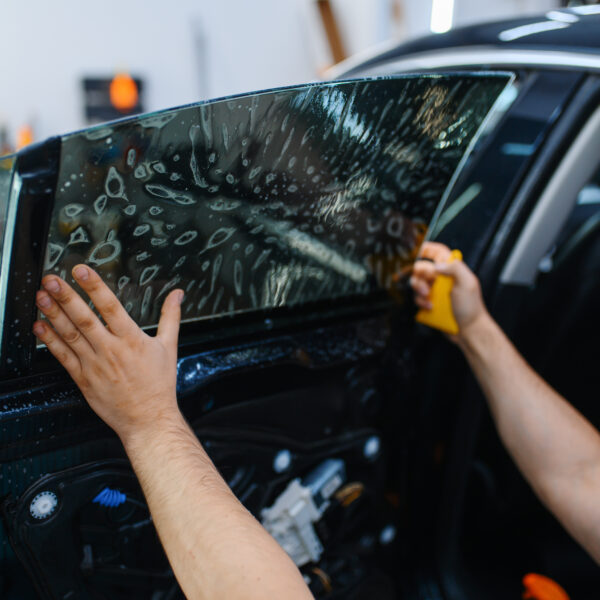 Male worker installs wetted car tinting, tuning service. Mechanic applying vinyl tint on vehicle window in garage, tinted automobile glass